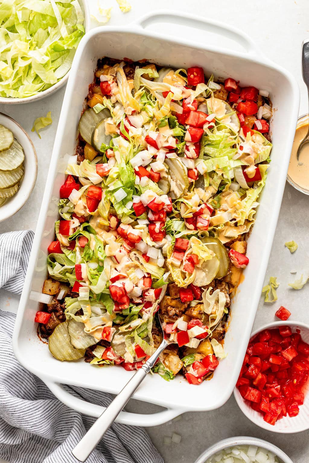 Rectangular white baking dish filled with a layered casserole topped with shredded lettuce, diced tomatoes, onions, cheese, and pickles, surrounded by bowls of the same ingredients.