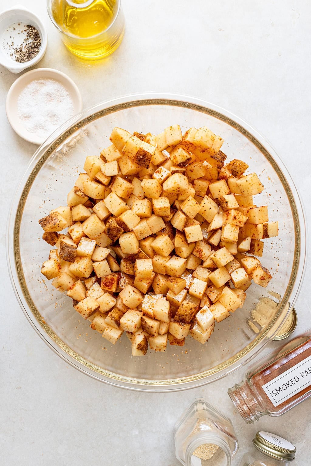 A glass bowl of diced potatoes seasoned with spices sits on a countertop next to salt, pepper, oil, smoked paprika, and garlic powder.