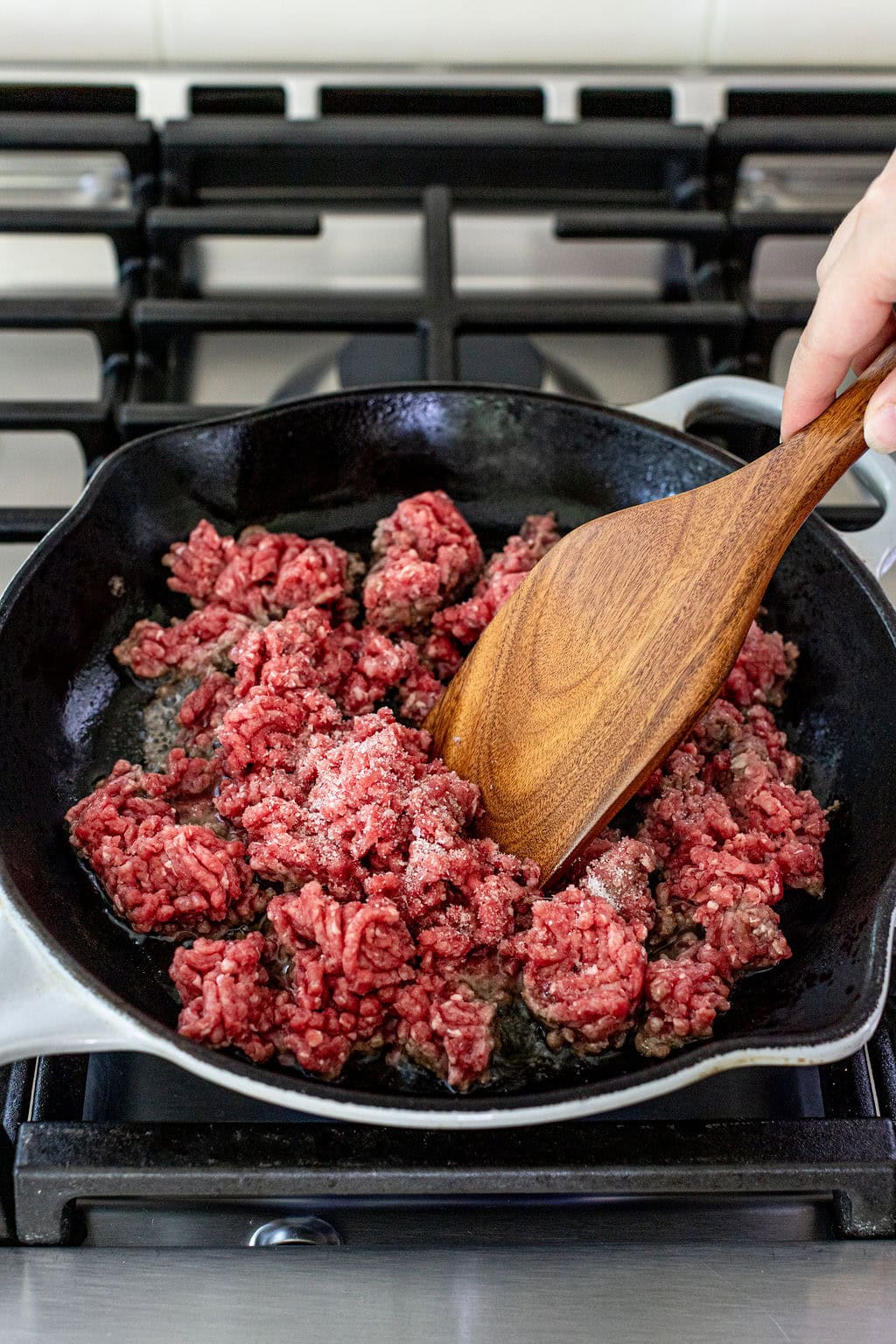 Ground beef is being cooked in a cast iron skillet on a stove, with a wooden spatula stirring the meat.