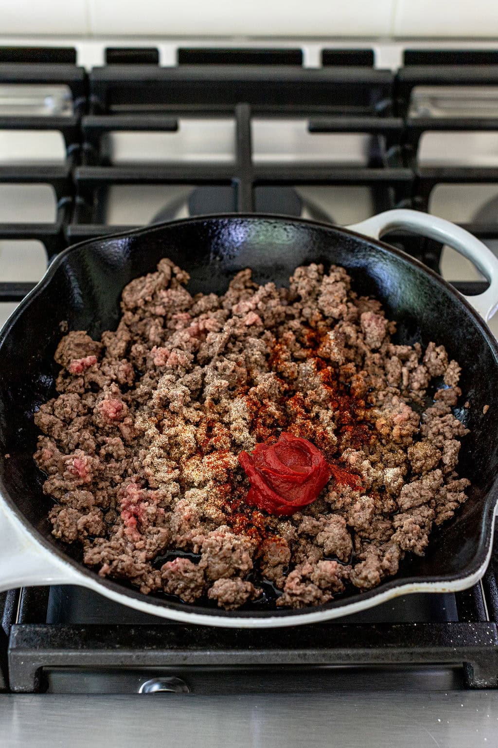 Ground beef cooking in a black skillet on a stove, with seasonings and a dollop of tomato paste added in the center.