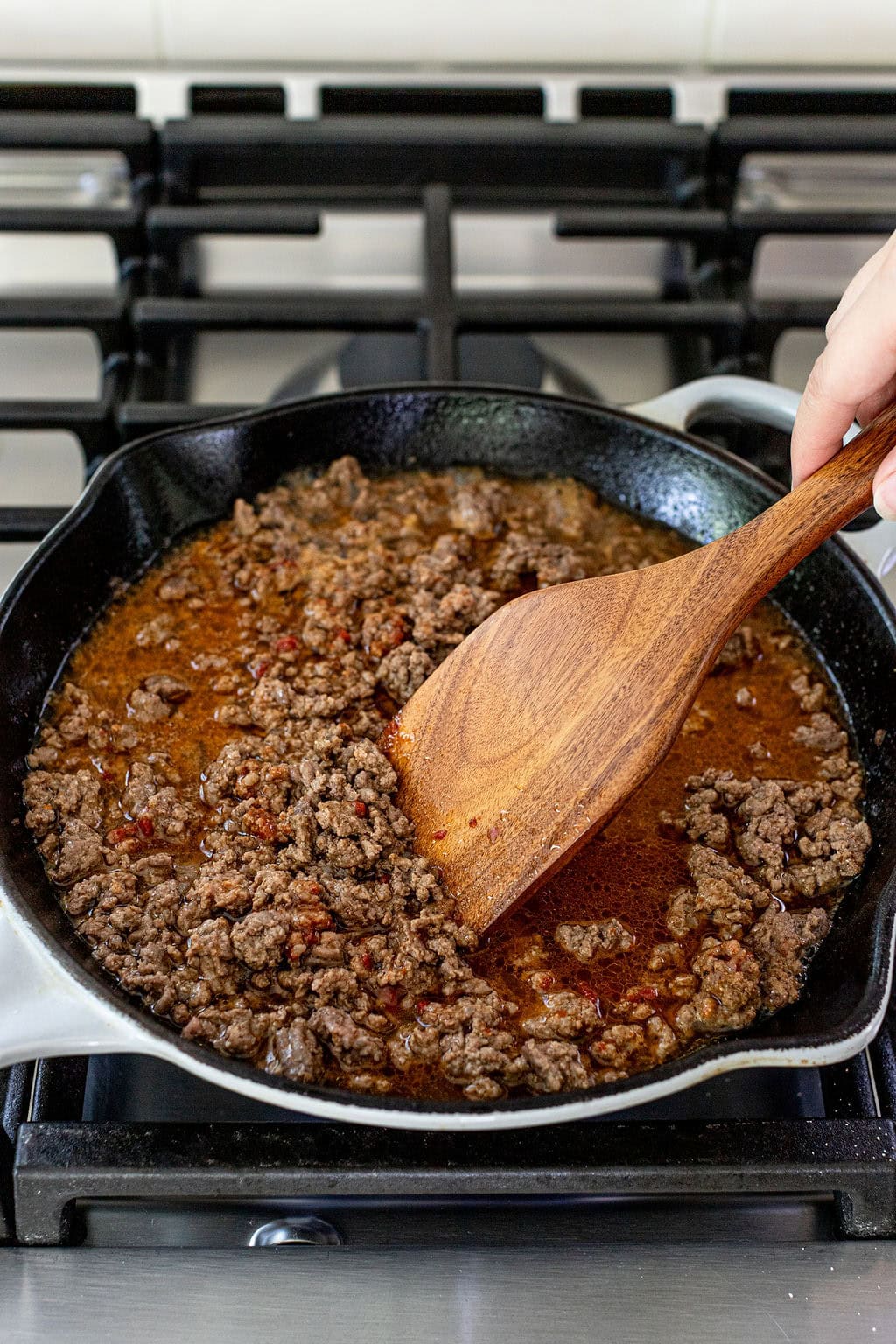 A hand uses a wooden spatula to stir seasoned ground beef cooking in a black cast iron skillet on a stove.