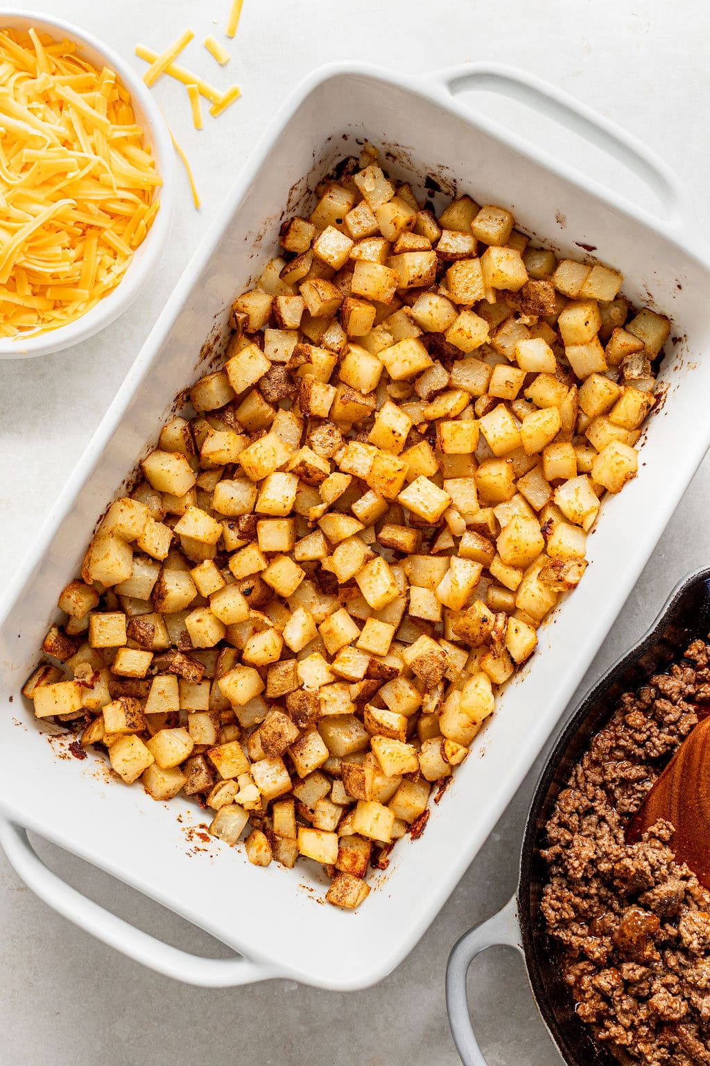 A white baking dish filled with roasted diced potatoes, next to a bowl of shredded cheddar cheese and a skillet of cooked ground beef.