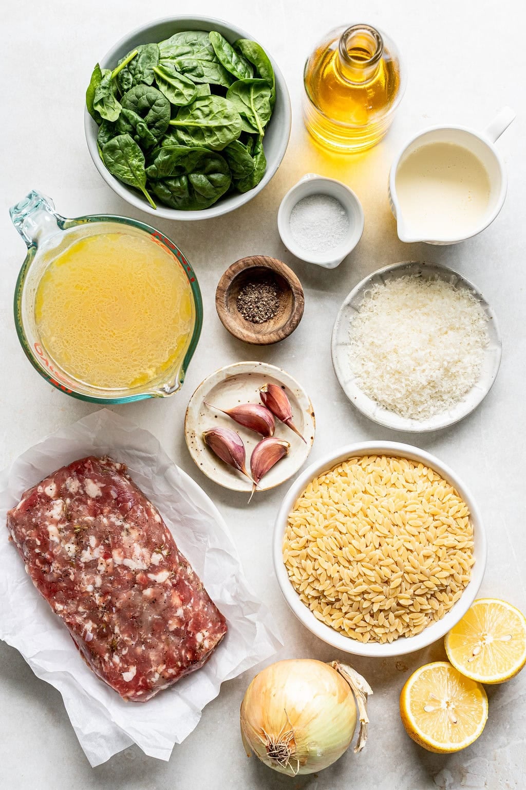 Ingredients on a countertop: spinach, olive oil, cream, chicken broth, salt, pepper, Parmesan, garlic, Italian sausage, orzo pasta, onion, and lemon halves.