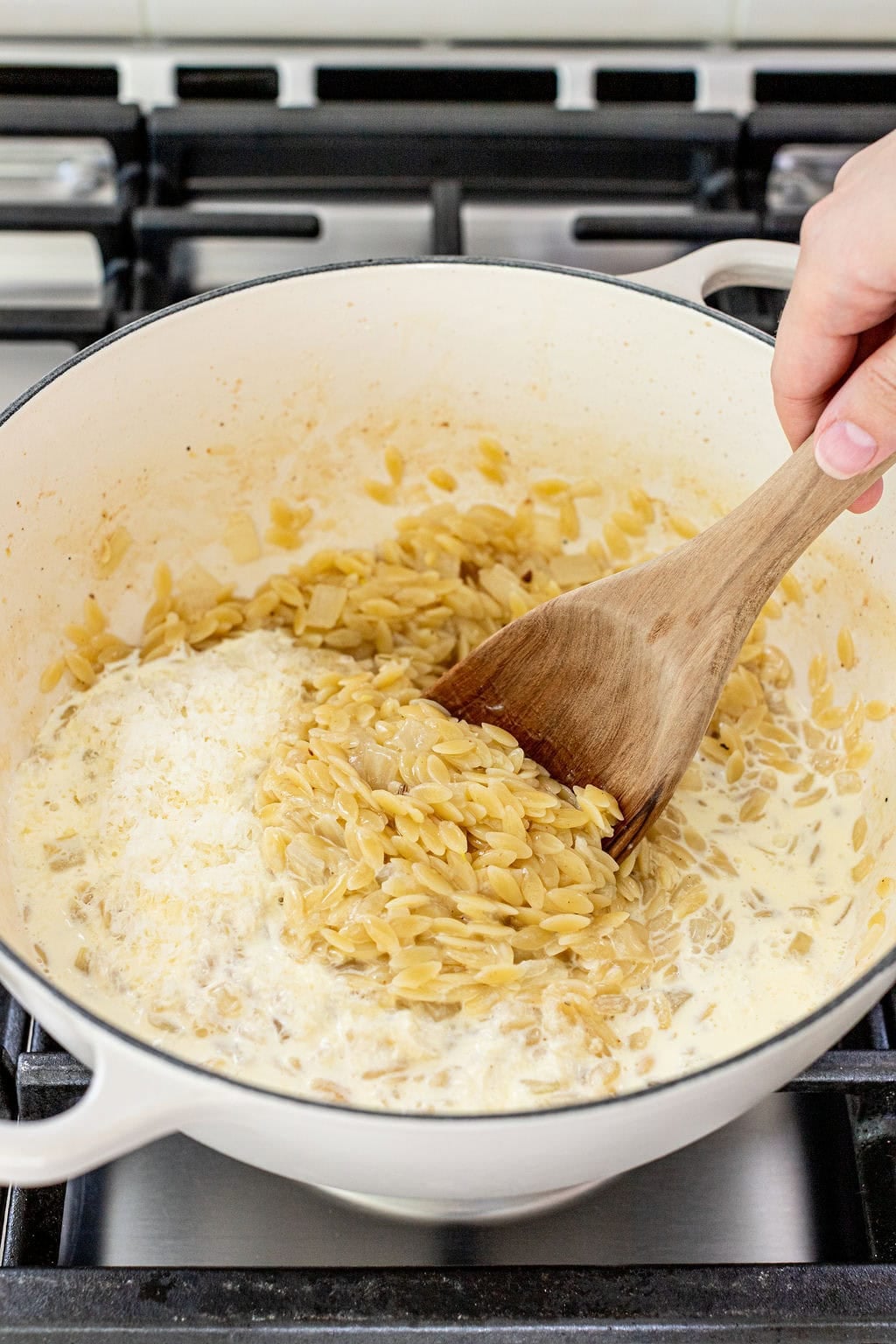 A hand stirs orzo pasta with a wooden spoon in a white pot on a stove, mixing it with cream.