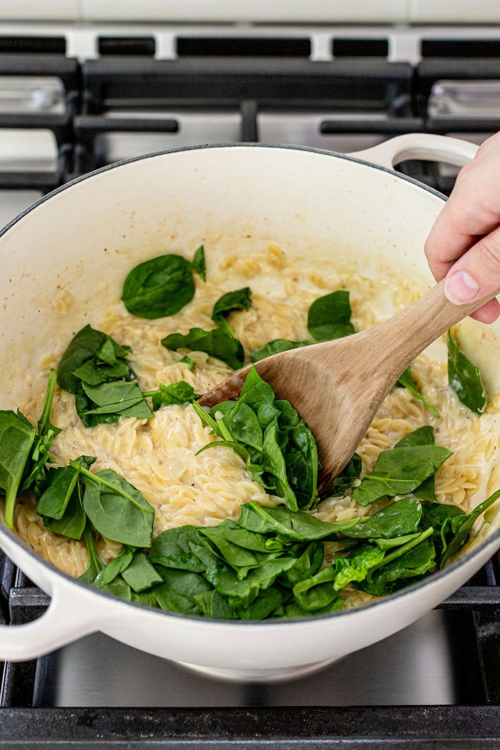 A hand stirs fresh spinach into creamy orzo pasta in a white pot on a stovetop.