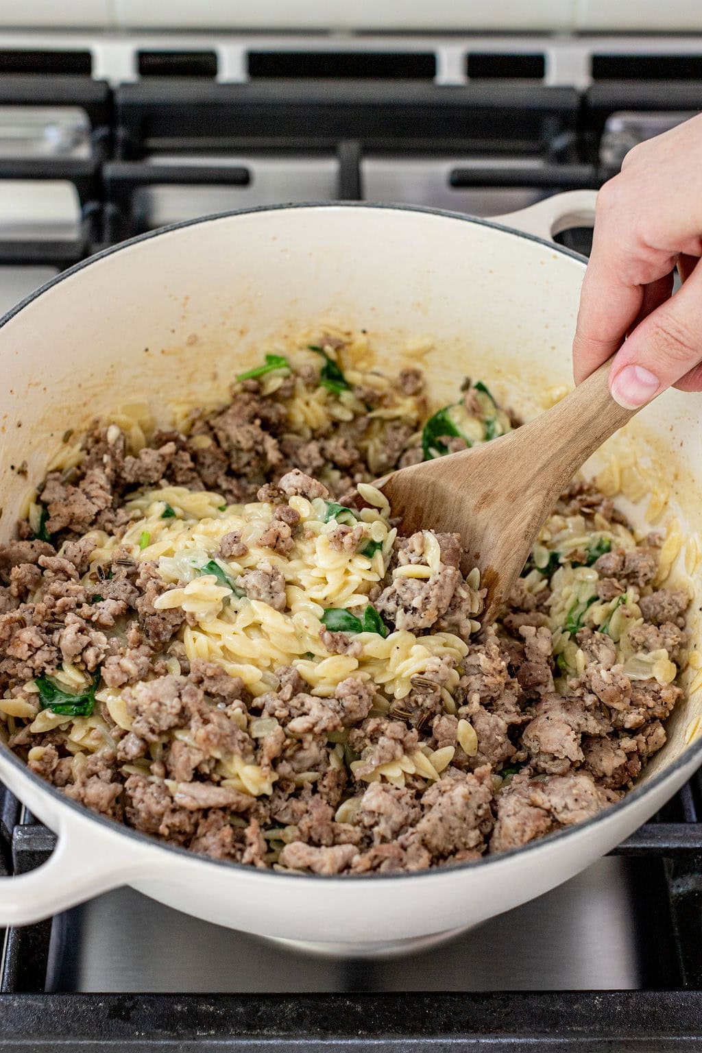 A hand stirs cooked ground meat, orzo pasta, and spinach in a large white pot on a stovetop.