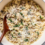 A pot of orzo pasta with ground meat, spinach, and grated cheese, mixed with a wooden spoon.