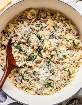 A pot of orzo pasta with ground meat, spinach, and grated cheese, mixed with a wooden spoon.