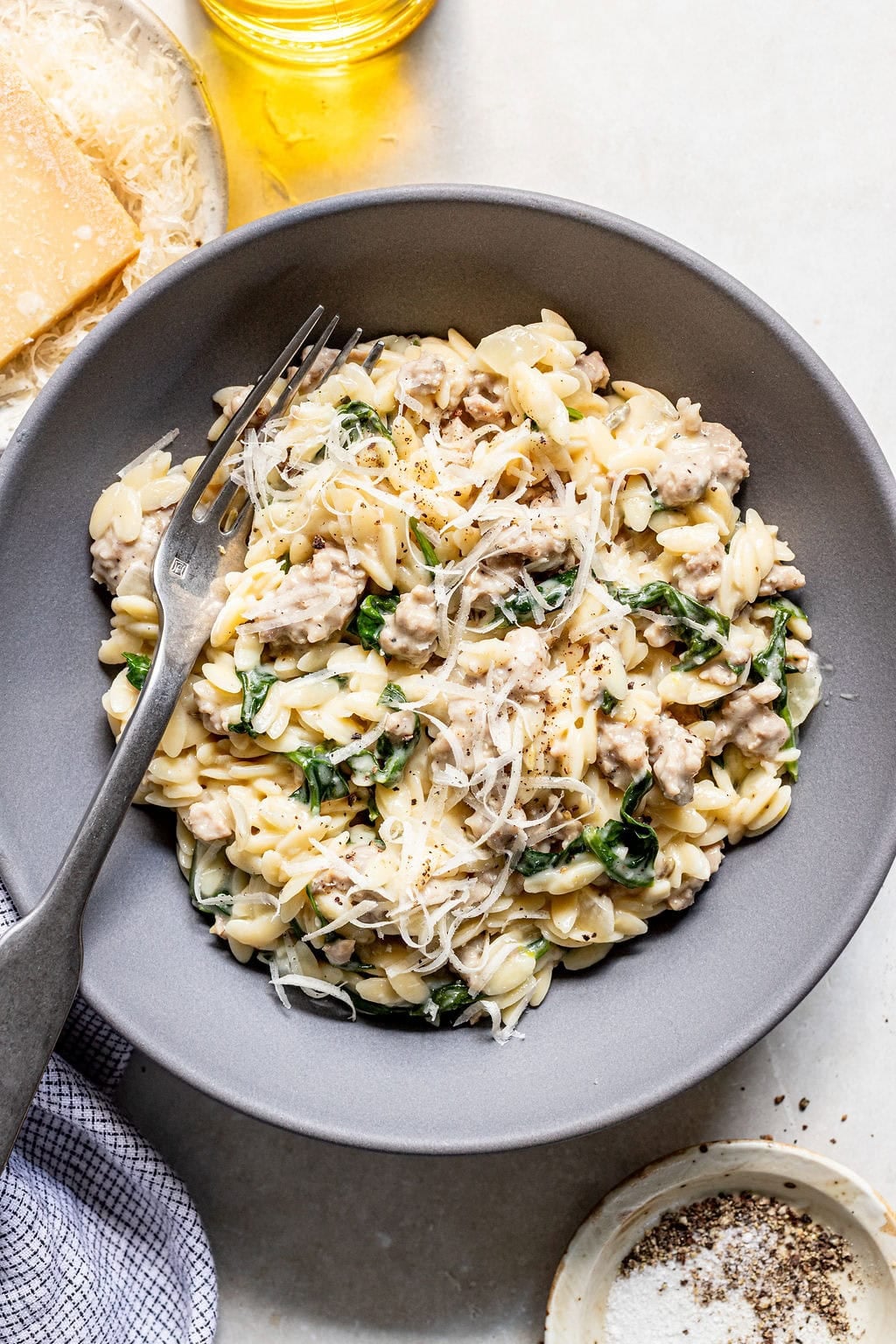 A bowl of orzo pasta with sausage, spinach, grated cheese, and black pepper, served with a fork. Parmesan, oil, and seasonings are on the side.