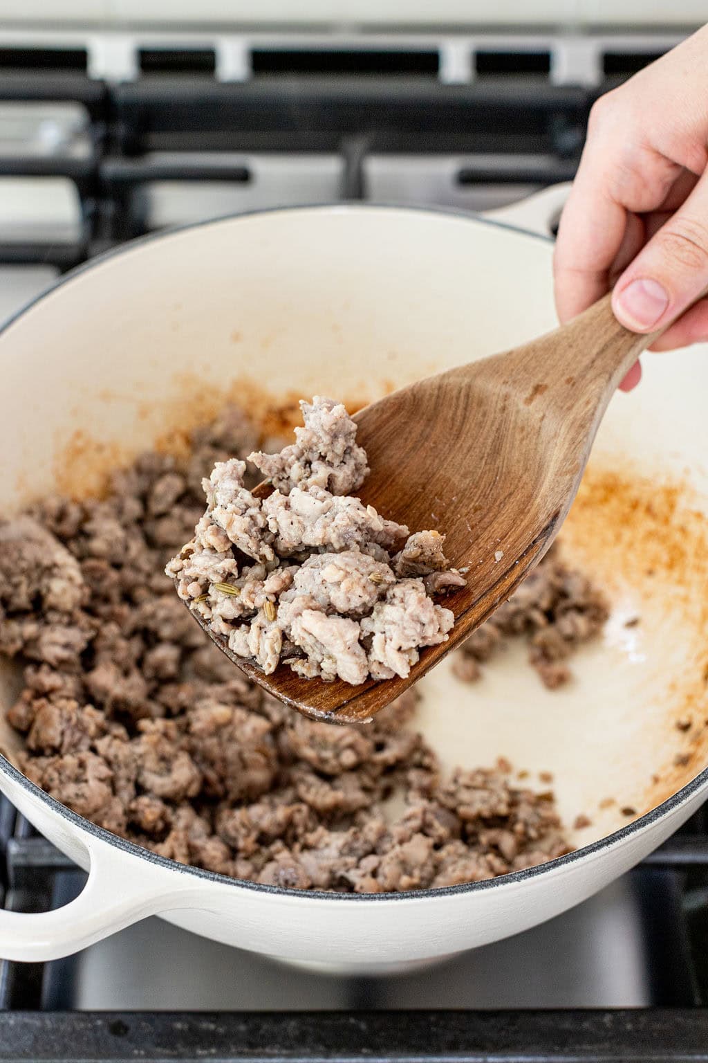 A hand holds a wooden spoon with cooked ground meat over a white pot on a stovetop.