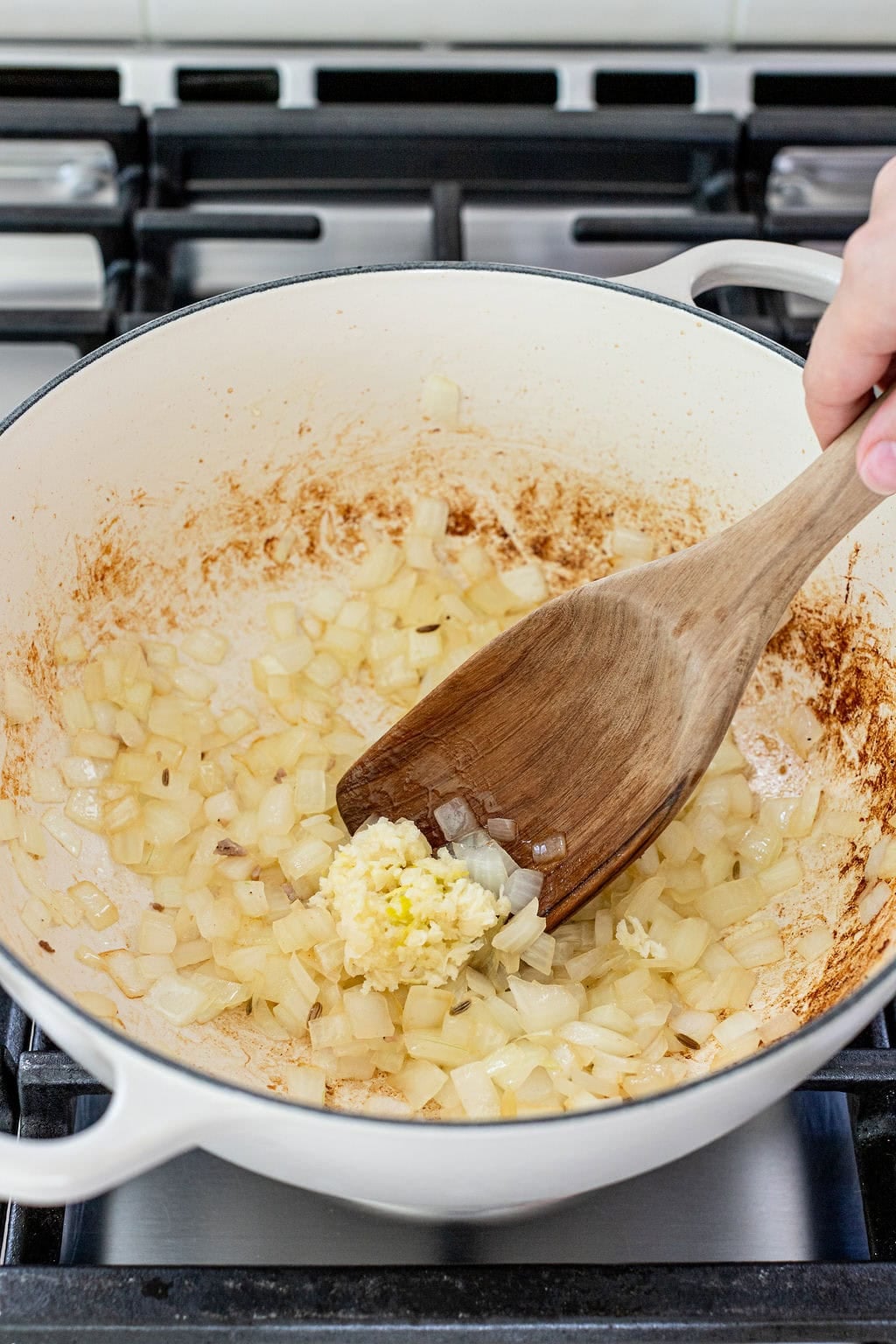 Diced onions and minced garlic being sautéed in a white pot on a stovetop, stirred with a wooden spatula.