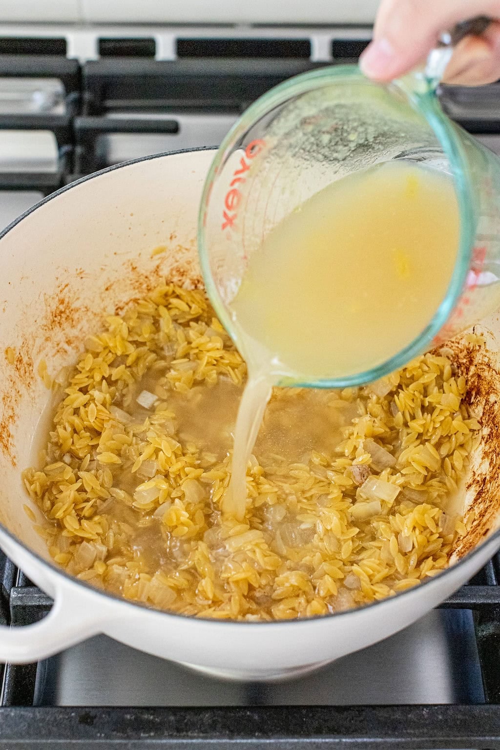 A hand pours broth from a glass measuring cup into a pot of sautéed orzo and onions on a stovetop.