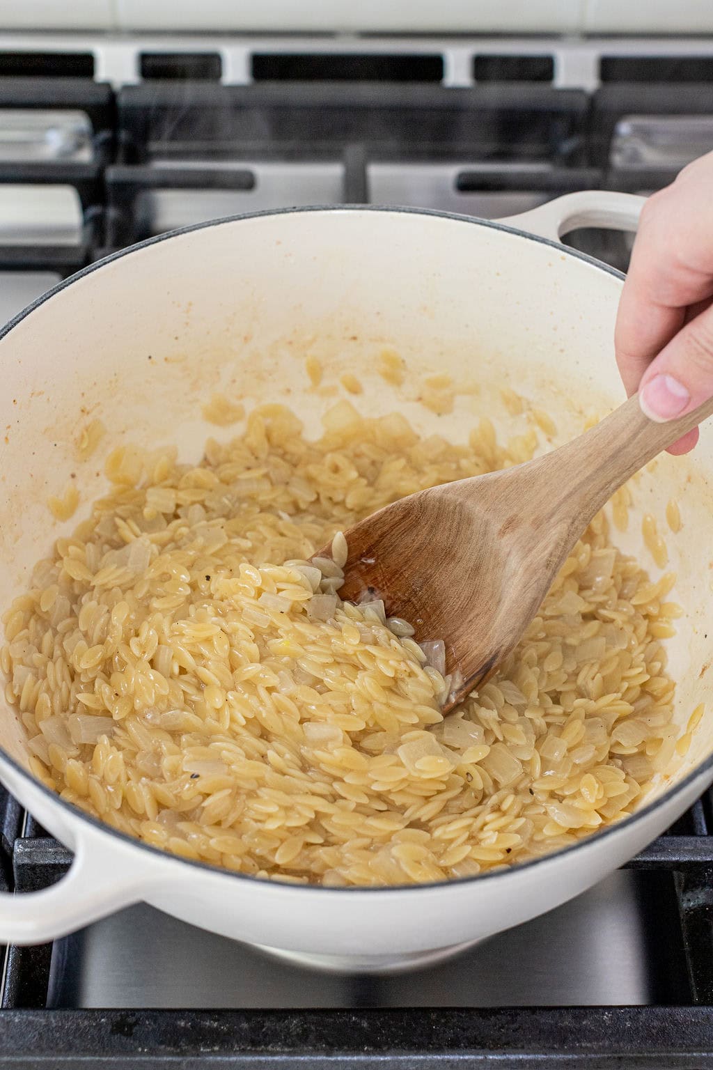 A hand stirs cooked orzo pasta in a white pot with a wooden spoon on a stovetop.