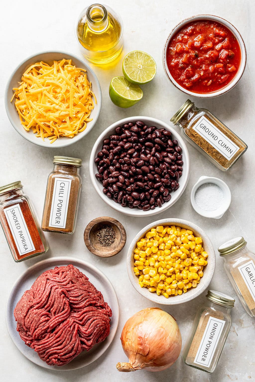Ingredients filled with ground beef, cheddar cheese, black beans, corn, onion, canned tomatoes, spices, pepper, lime, and olive oil arranged on a white surface.
