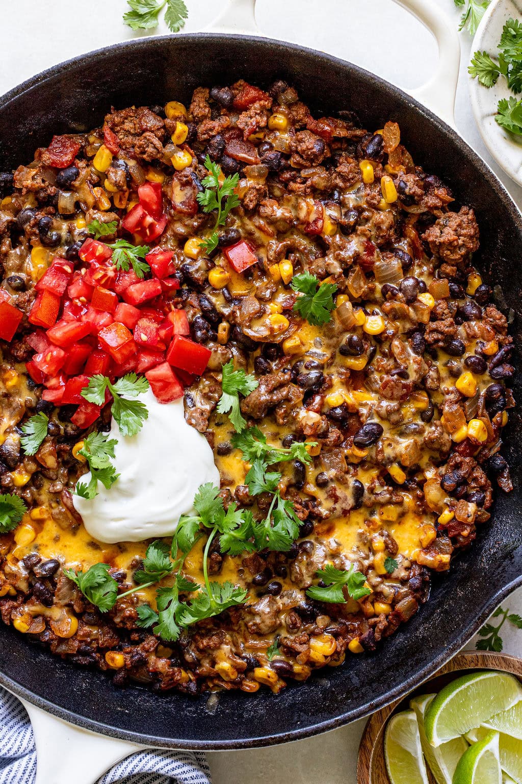 A skillet filled with cooked ground beef, black beans, corn, melted cheese, chopped tomatoes, cilantro, and a dollop of sour cream.