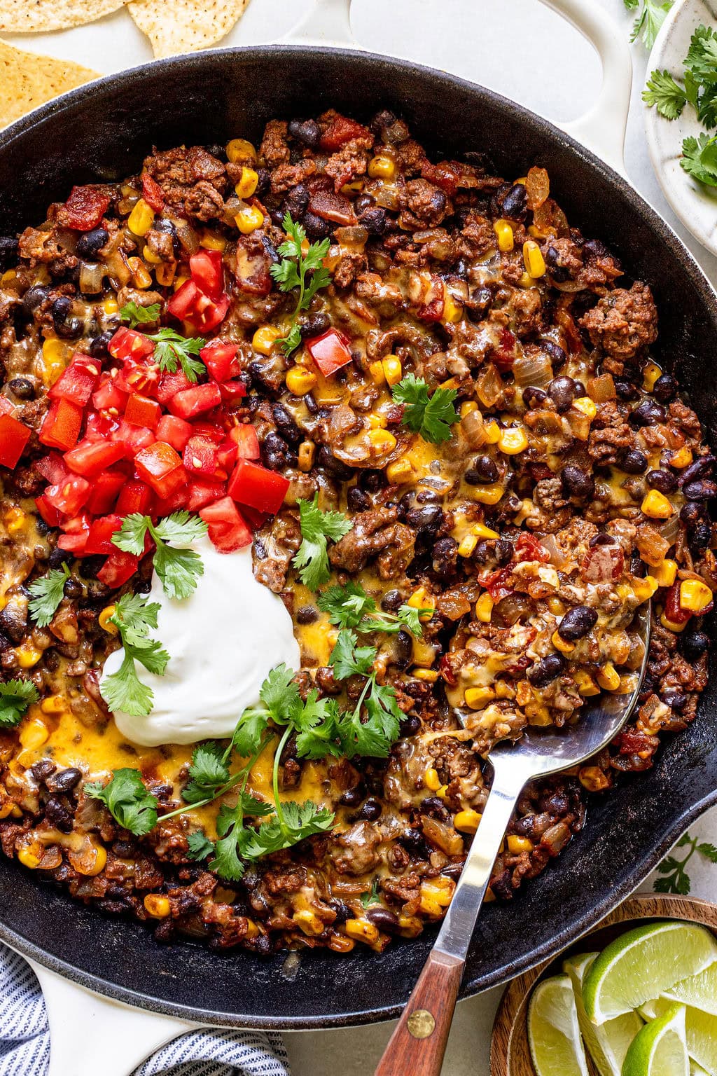 Skillet filled with ground beef, black beans, corn, melted cheese, diced tomatoes, fresh cilantro, and a dollop of sour cream, with a spoon and lime wedges on the side.