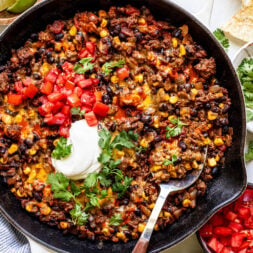 A skillet filled with cooked ground beef, black beans, corn, melted cheese, and tomatoes, topped with sour cream and cilantro, with a serving spoon and garnished with avocado, lime, and tortilla chips.