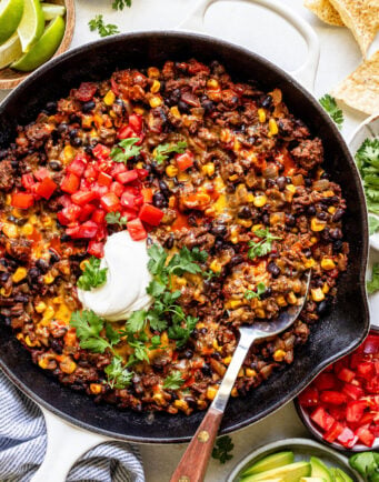 A skillet filled with cooked ground beef, black beans, corn, melted cheese, and tomatoes, topped with sour cream and cilantro, with a serving spoon and garnished with avocado, lime, and tortilla chips.