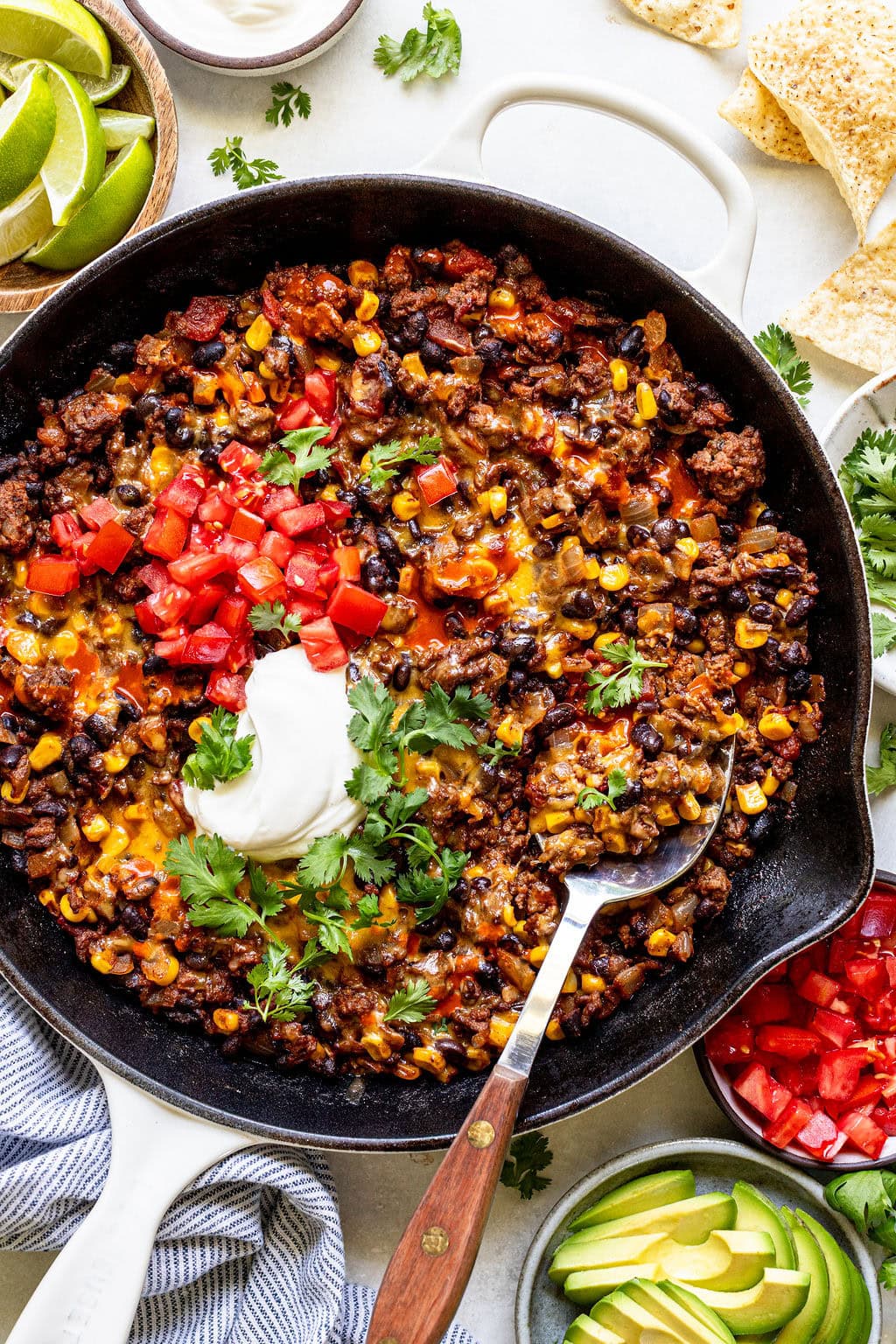 A skillet filled with cooked ground beef, black beans, corn, melted cheese, and tomatoes, topped with sour cream and cilantro, with a serving spoon and garnished with avocado, lime, and tortilla chips.