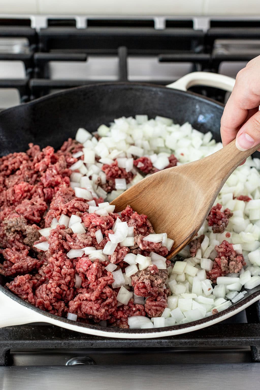 Ground beef and diced onions being stirred with a wooden spatula in a skillet on a stove.