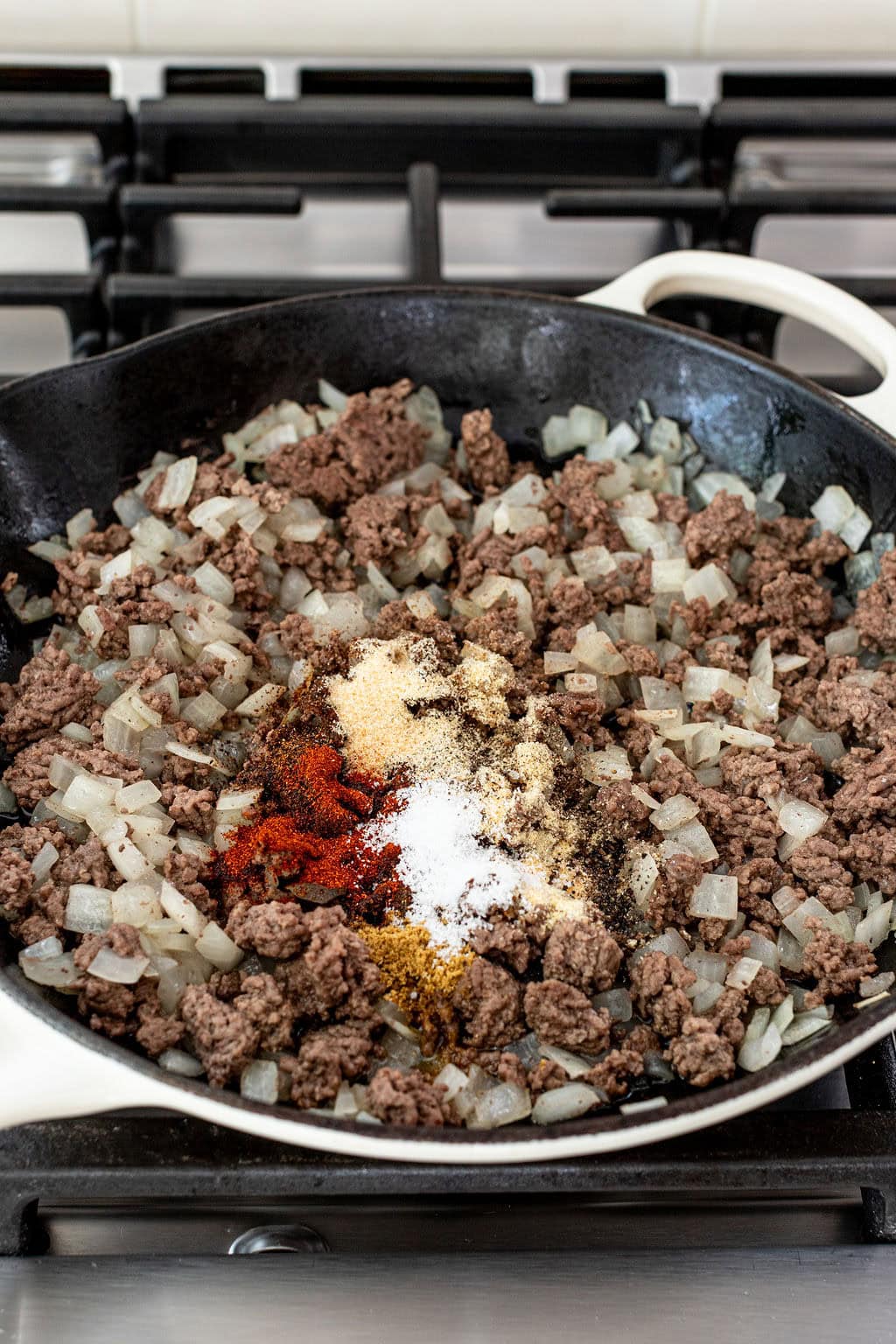 Ground beef and chopped onions cooking in a skillet on a stove, with various spices sprinkled on top, ready to be mixed.