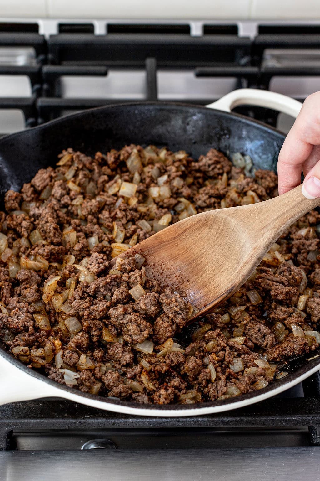 Cooked ground beef and diced onions being stirred in a skillet on a stove with a wooden spoon.