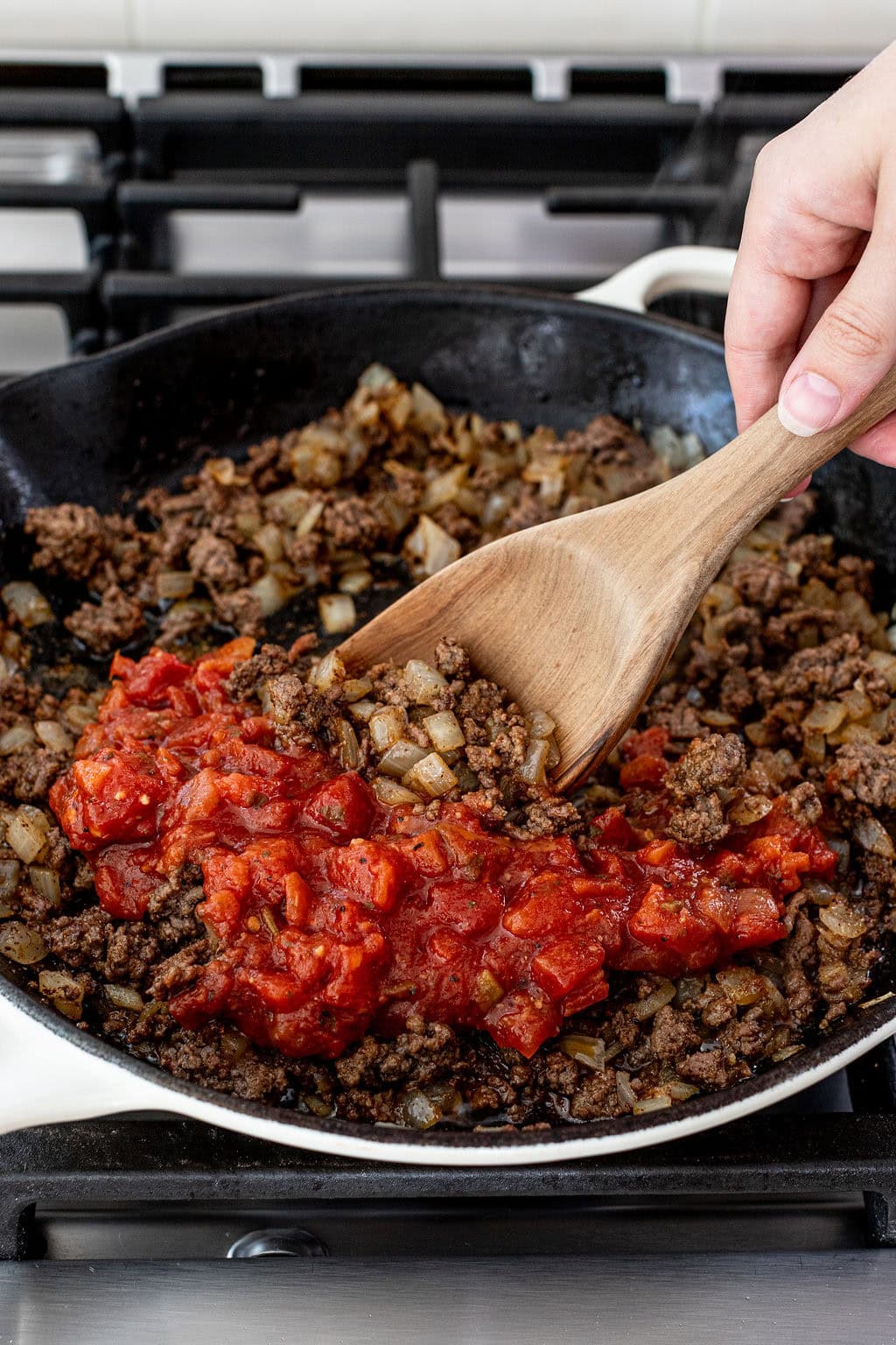 Ground beef, chopped onions, and diced tomatoes are being stirred together in a skillet on a stovetop using a wooden spoon.