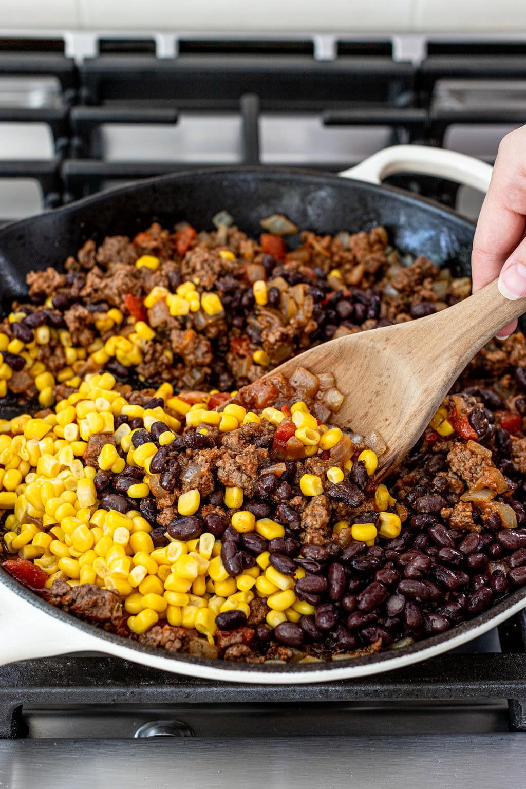 A skillet on a stovetop filled with cooked ground beef, black beans, corn, and diced vegetables being stirred with a wooden spoon.