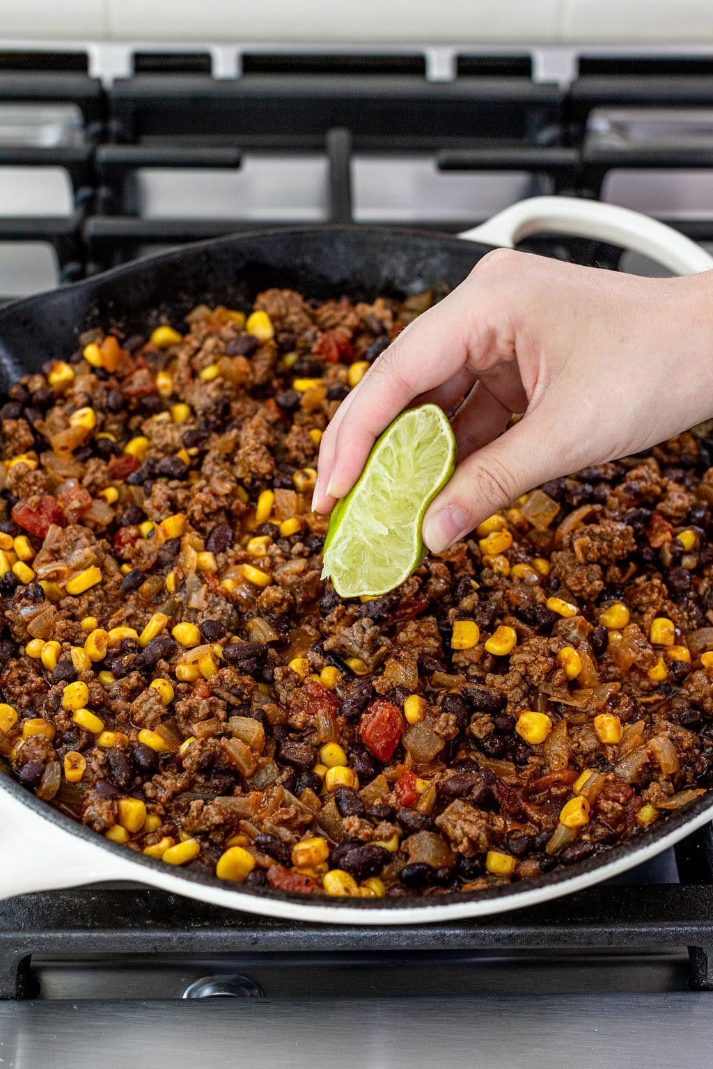 A hand squeezes a lime wedge over a skillet filled with a cooked mixture of ground beef, corn, black beans, tomatoes, and onions on a stovetop.