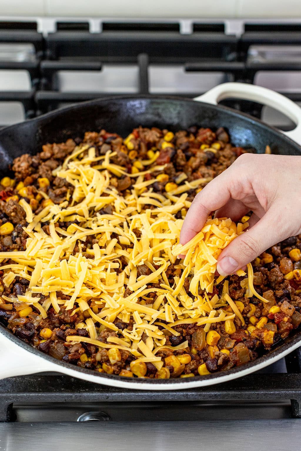 A hand sprinkles shredded cheddar cheese over a skillet filled with cooked ground beef, corn, and black beans on a stove.
