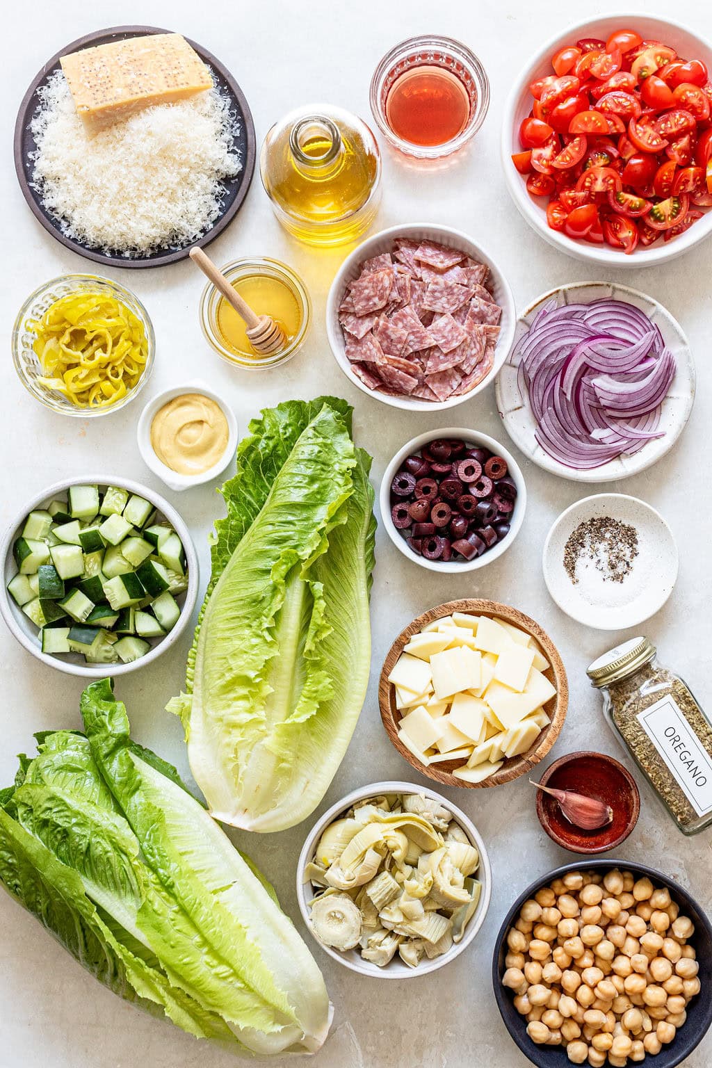 Top-down view of salad ingredients including romaine lettuce, cherry tomatoes, cucumber, olives, cheese, chickpeas, salami, artichokes, red onion, pepperoncini, oil, vinegar, honey, and seasonings.