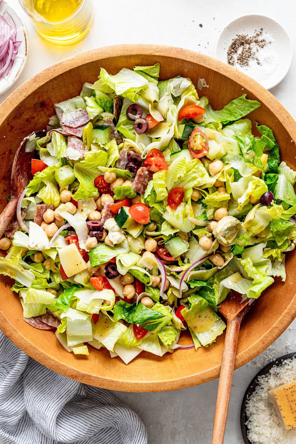 A wooden bowl of chopped salad with lettuce, cherry tomatoes, red onion, chickpeas, olives, artichoke hearts, cheese, and a wooden spoon, with oil and pepper nearby.