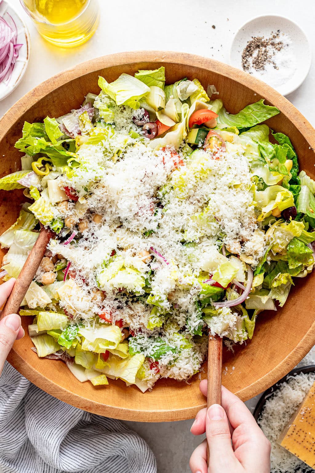 A large wooden bowl of salad with lettuce, tomatoes, onions, olives, and grated cheese, being tossed with wooden utensils.