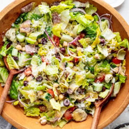 Large wooden bowl of chopped salad with lettuce, tomatoes, olives, chickpeas, artichokes, red onion, and grated cheese, served with wooden utensils. A small dish of pepper and oil is nearby.