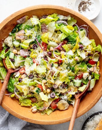 Large wooden bowl of chopped salad with lettuce, tomatoes, olives, chickpeas, artichokes, red onion, and grated cheese, served with wooden utensils. A small dish of pepper and oil is nearby.