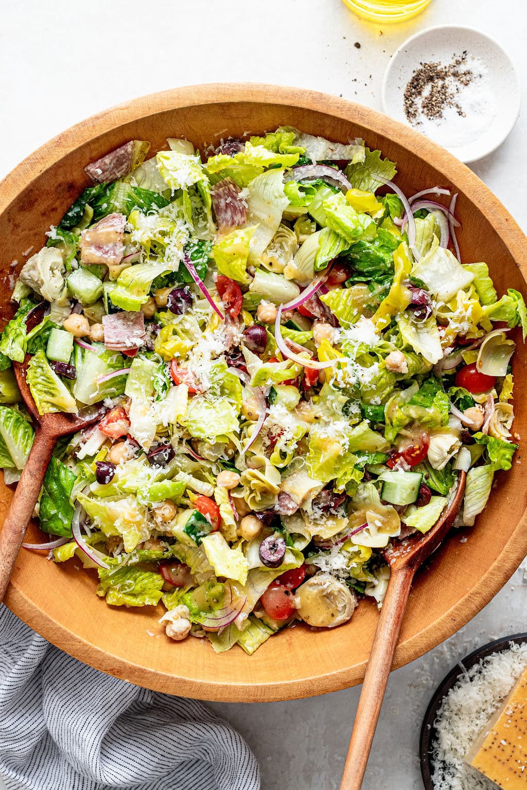Large wooden bowl of chopped salad with lettuce, tomatoes, olives, chickpeas, artichokes, red onion, and grated cheese, served with wooden utensils. A small dish of pepper and oil is nearby.