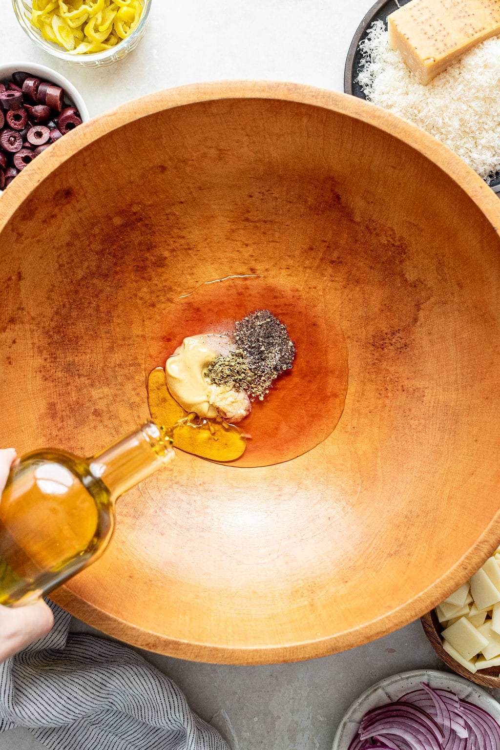 A hand pours olive oil into a large wooden bowl containing mustard and seasonings, with bowls of salad ingredients visible around the edges.