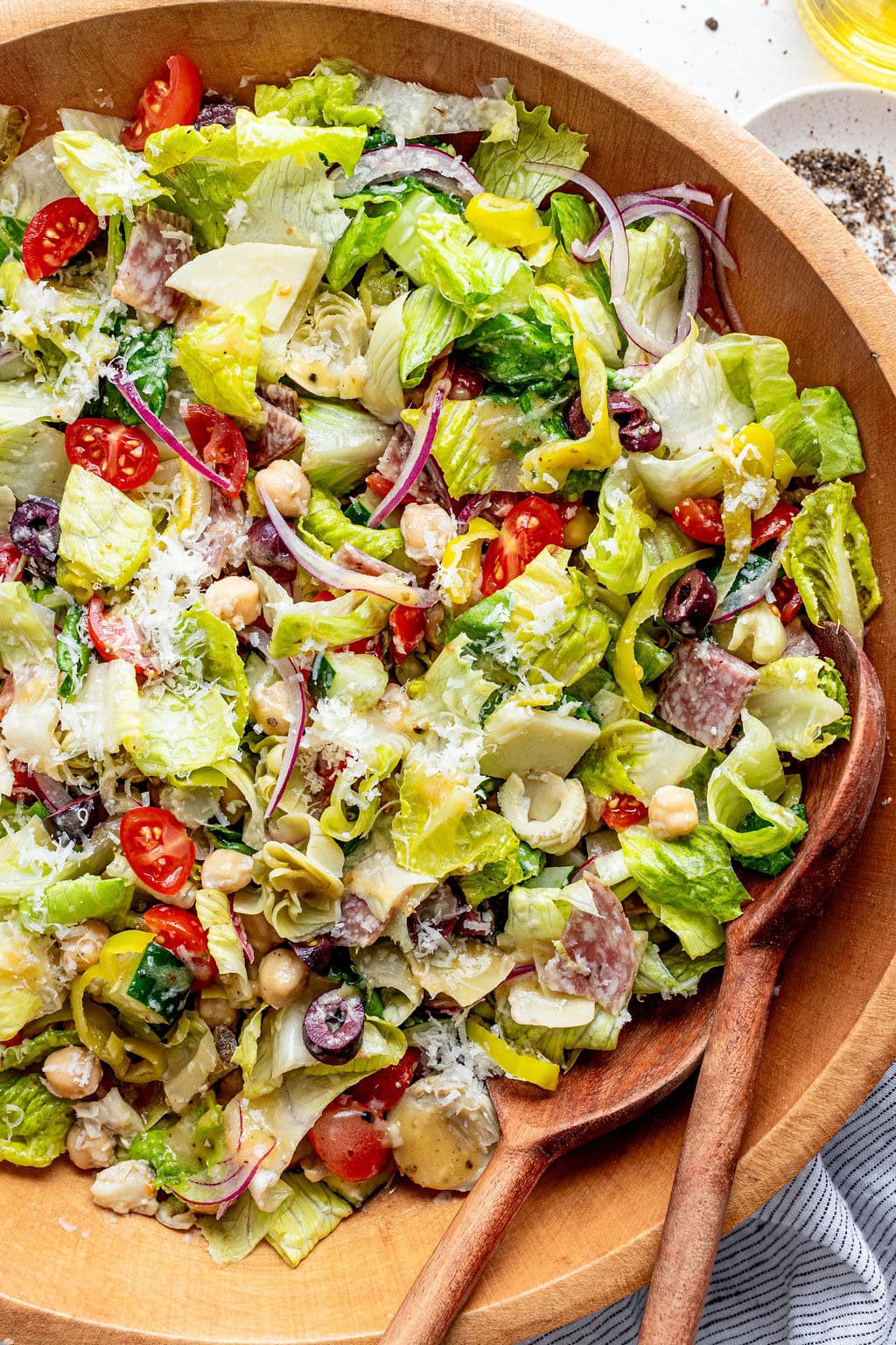 A wooden bowl filled with chopped salad including lettuce, cherry tomatoes, red onion, olives, chickpeas, pepperoncini, artichoke hearts, salami slices, and grated cheese, with wooden salad servers.