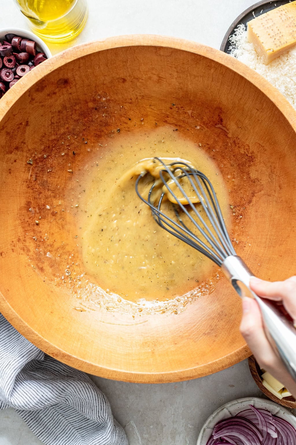A hand uses a whisk to mix dressing in a wooden bowl. Surrounding the bowl are ingredients like sliced olives, grated cheese, rice, and red onion.