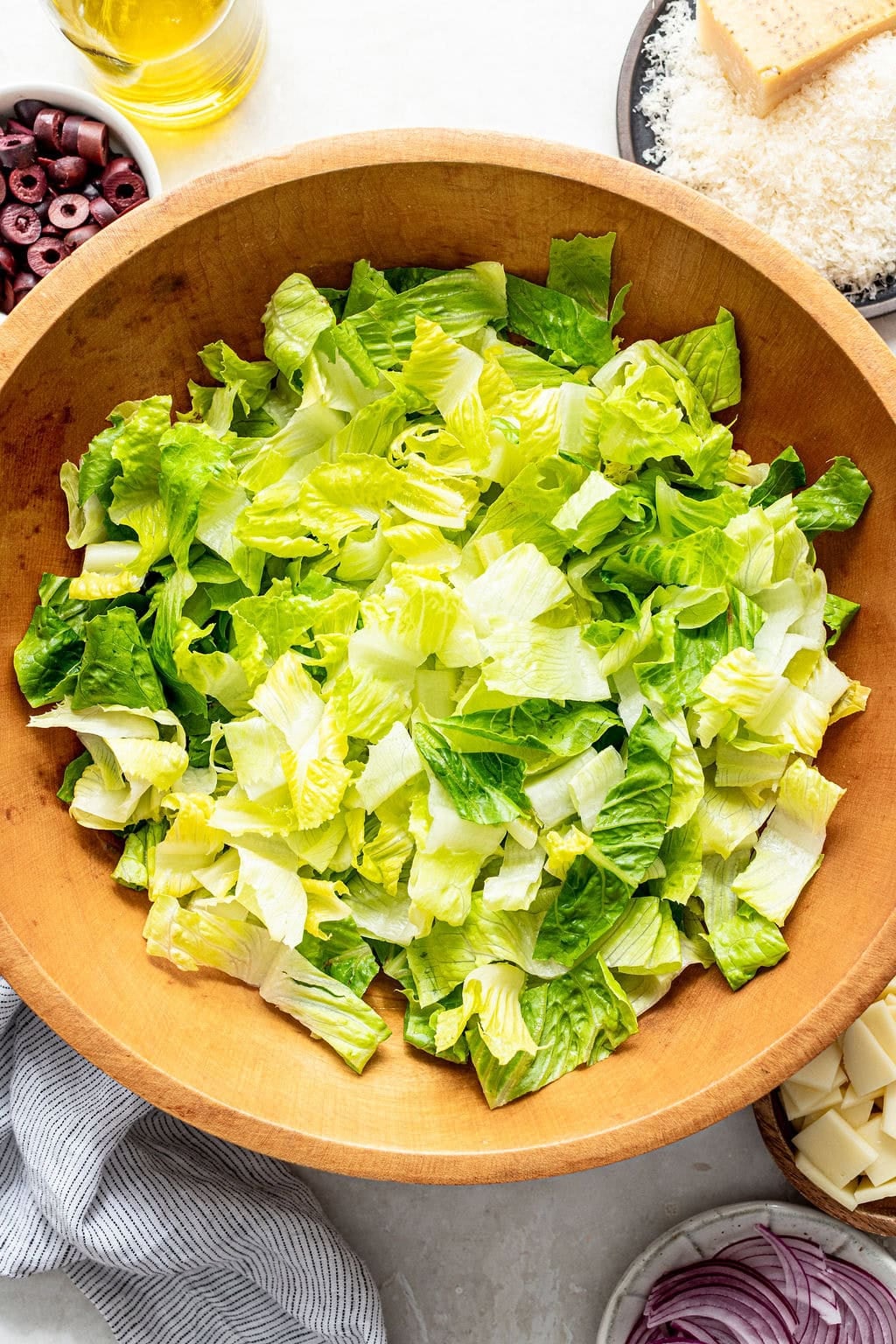 A wooden bowl filled with chopped romaine lettuce sits on a countertop, surrounded by bowls of olives, sliced onions, grated cheese, and a bottle of olive oil.