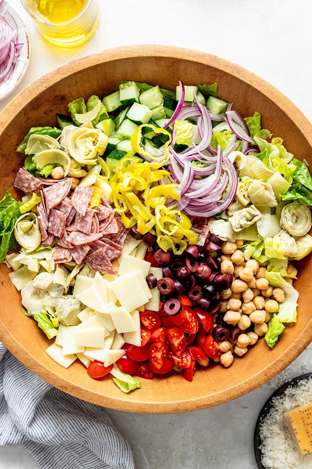 A wooden bowl filled with chopped lettuce, cucumbers, red onion, artichokes, salami, mozzarella, tomatoes, olives, chickpeas, and pepperoncini.