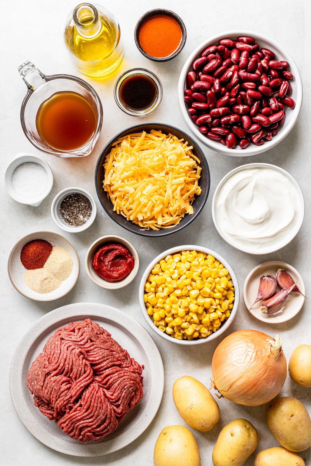 Assorted chili ingredients on a white surface, including ground beef, potatoes, onion, garlic, corn, kidney beans, cheese, sour cream, broth, oil, spices, tomato paste, and sauces.