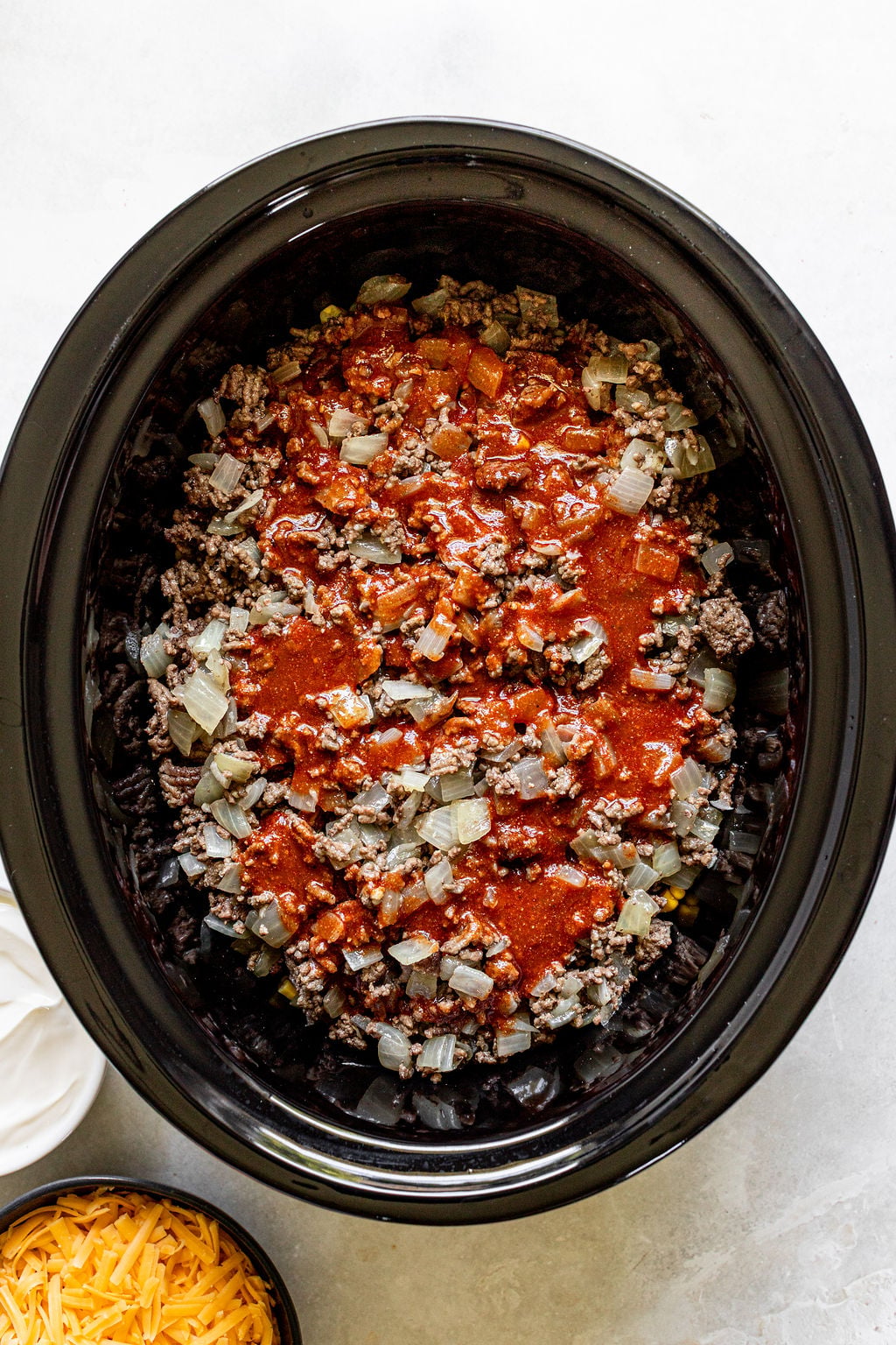 A slow cooker filled with cooked ground beef, diced onions, and tomato sauce, with bowls of shredded cheese and sour cream nearby.