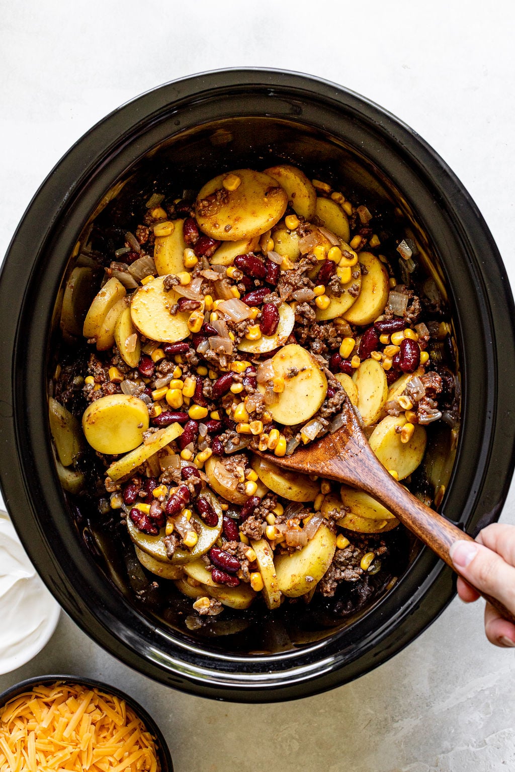 A hand stirs sliced potatoes, ground meat, corn, beans, and onions in a black slow cooker. Bowls of sour cream and shredded cheese sit nearby.