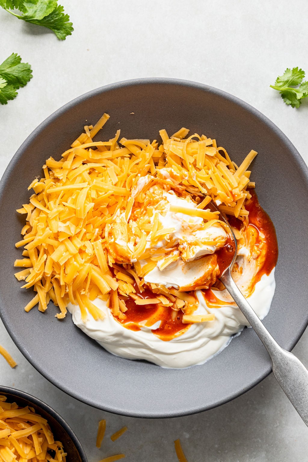A gray bowl with shredded cheddar cheese, sour cream, and hot sauce being mixed with a spoon.