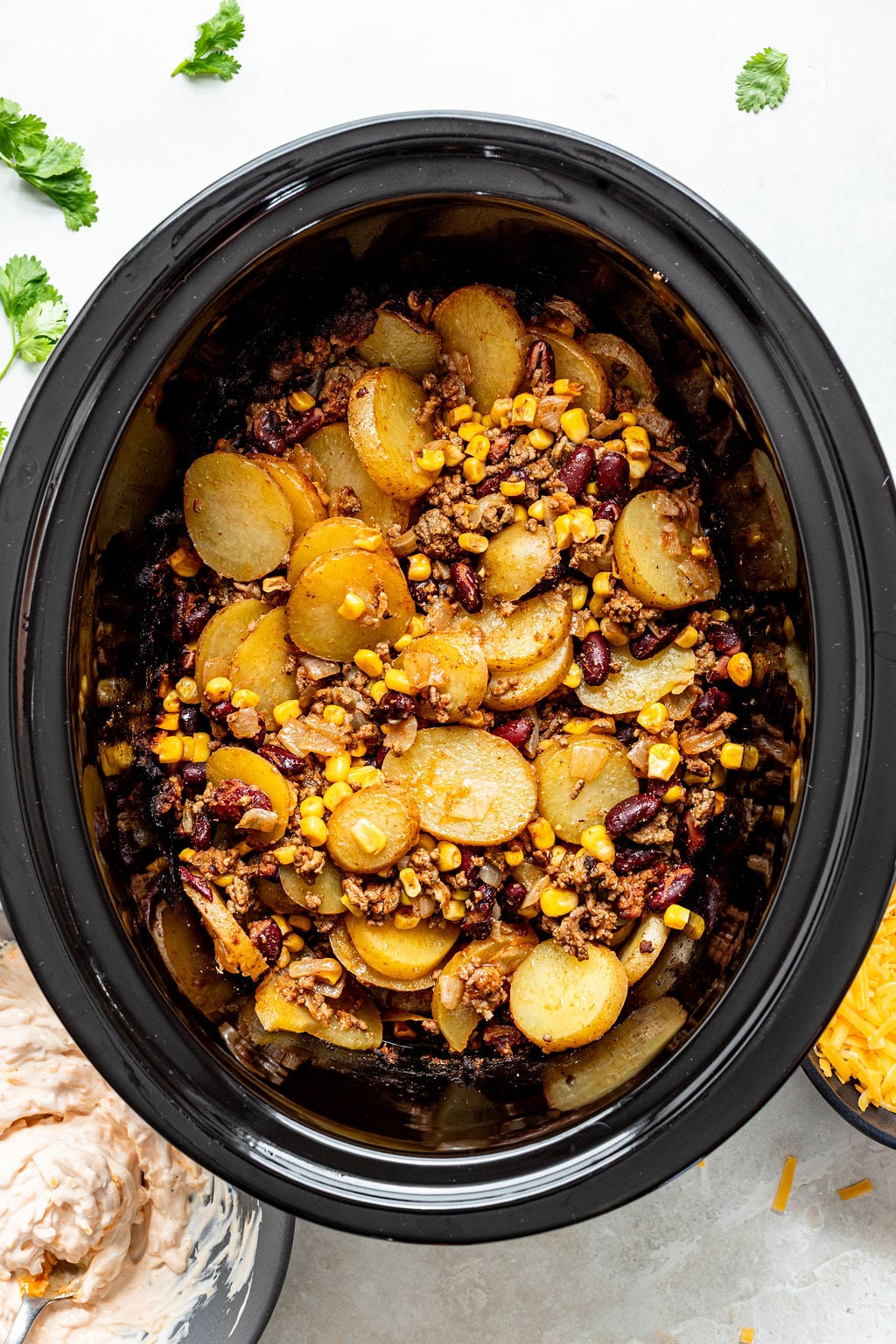 A black slow cooker filled with sliced potatoes, ground meat, corn, beans, and onions, with cilantro and shredded cheese on the side.