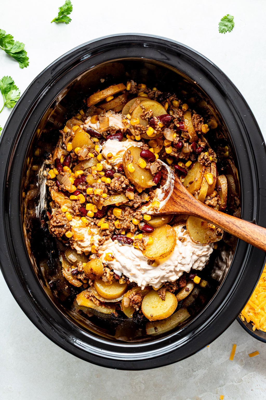 Slow cooker filled with layers of ground meat, corn, sliced potatoes, beans, and a dollop of cream mixture, with a wooden spoon resting inside.