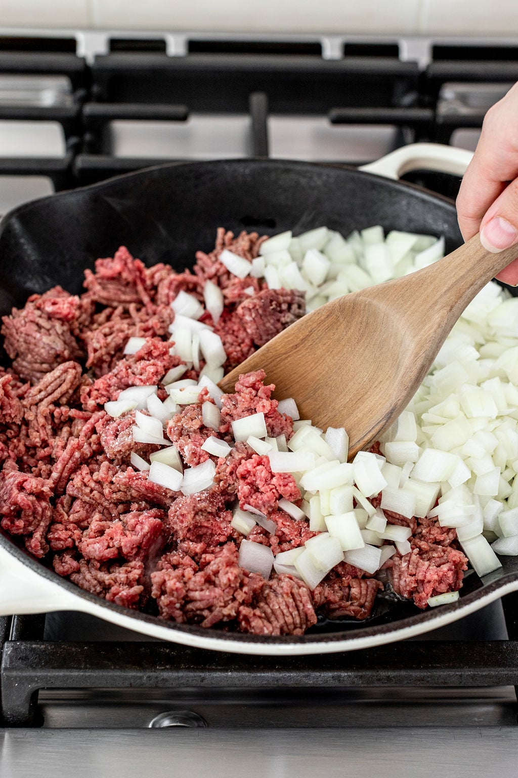 Ground beef and chopped onions are being cooked together in a black skillet on a stovetop, stirred with a wooden spatula.