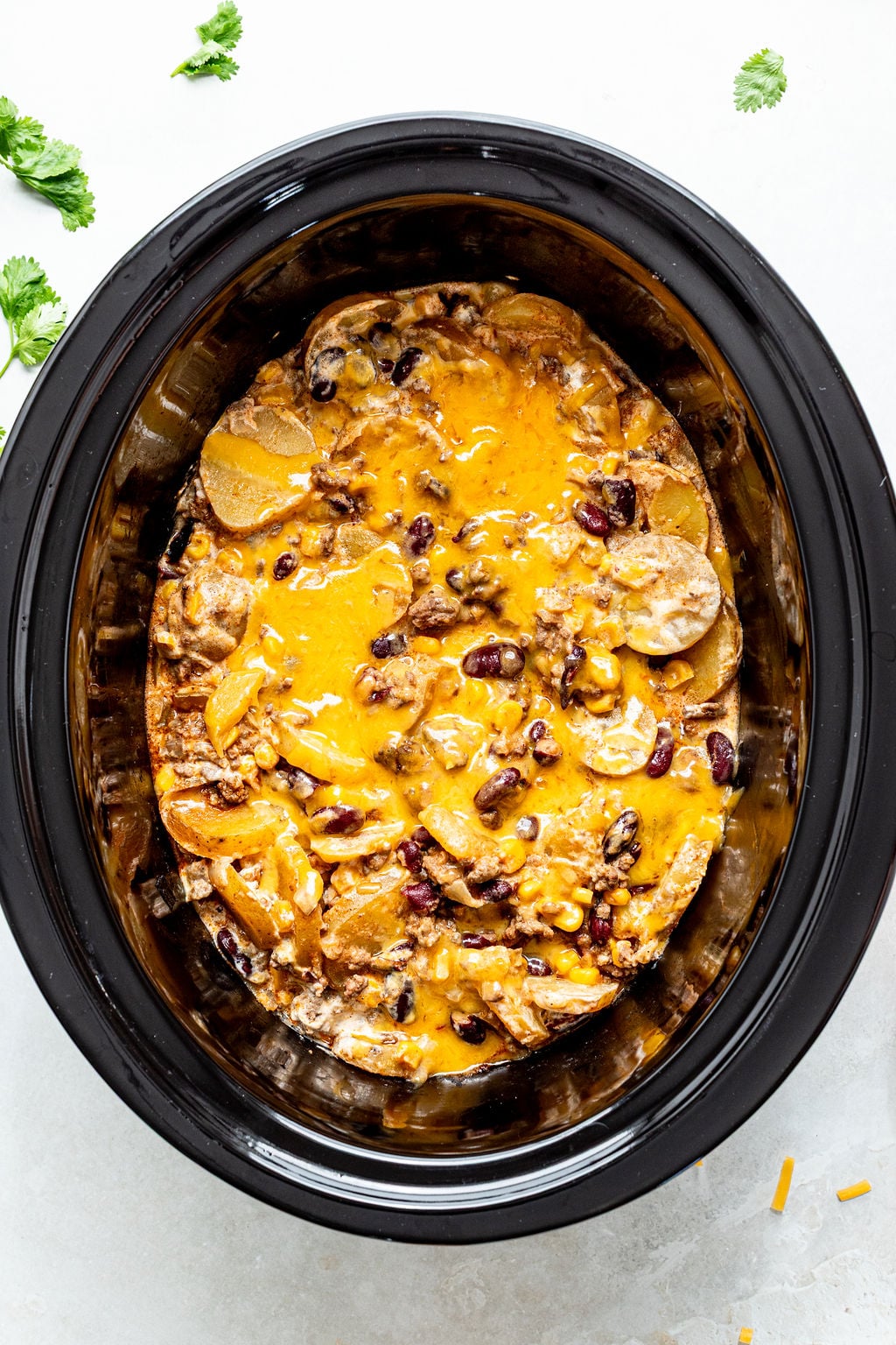 A slow cooker filled with a baked cheesy casserole containing beans, potato slices, and ground meat, garnished with melted cheese on top.