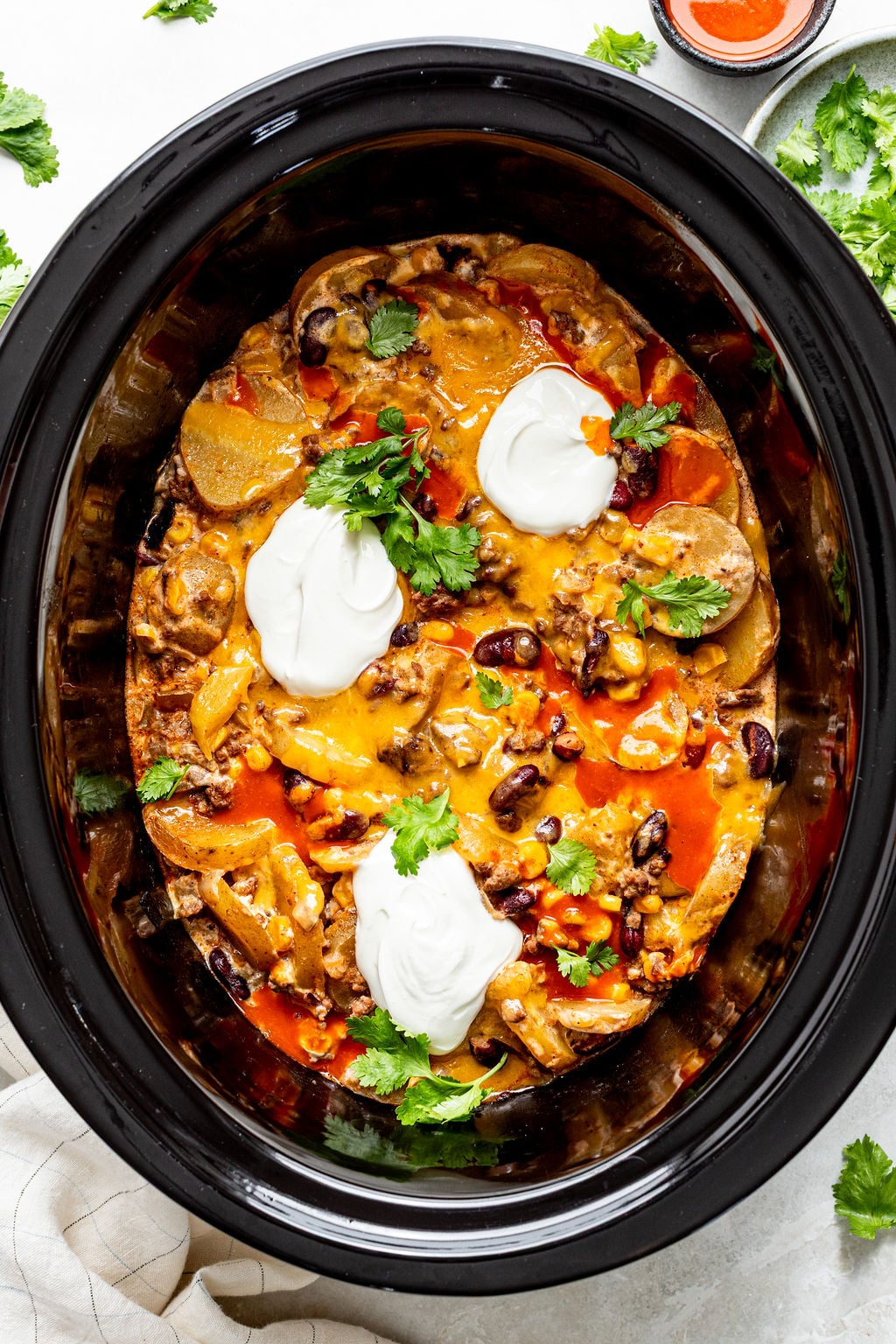 A slow cooker filled with cheesy casserole topped with sour cream and fresh cilantro, featuring visible beans, potatoes, and vegetables.